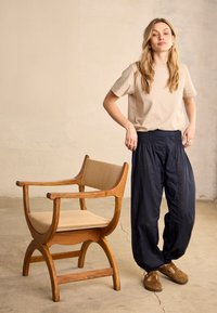 Kvinde med langt blondt hår står ved siden af en træstol med beige puder, iført en beige skjorte, løse sorte bukser og brune sko.