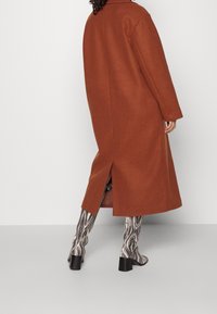 Abrigo largo de color óxido con corte recto, aberturas laterales y un cuello con solapa, combinado con botas altas hasta la rodilla de patrón en espiral en blanco y negro.