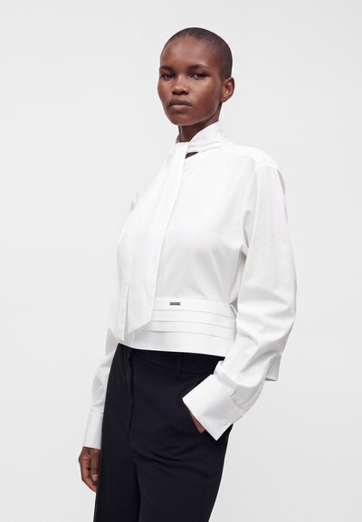 Femme aux cheveux courts porte un chemisier blanc à manches longues avec un col montant et un nœud, ainsi qu’un pantalon noir, debout devant un fond clair uni.