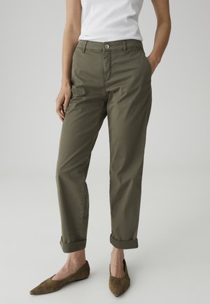 Vrouw met olijfgroene omslagbroek, wit mouwloos topje en bruine puntige platte schoenen, staand met één hand in de zak.