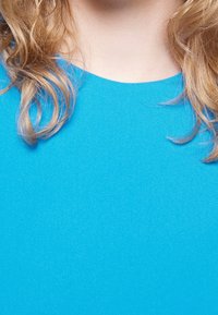 Jasně modrá látka s hladkým povrchem, s kulatým výstřihem. Vlnité blond vlasy částečně viditelné na pozadí z látky.
