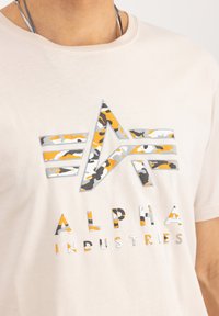 Beige tričko s šedým, oranžovým a maskáčovým hvězdným logem nad textem "ALPHA INDUSTRIES" v různých barvách a stylech.