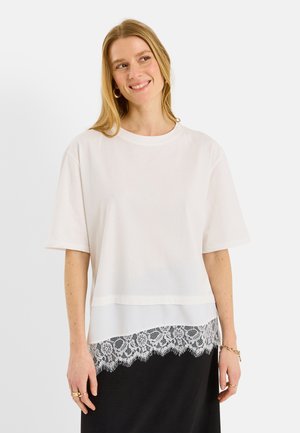 Mujer sonriente con una blusa blanca de manga corta y holgada con ribete de encaje sobre una falda negra, pendientes de aro dorados, anillos y pulsera.