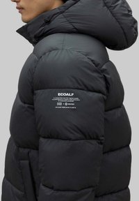 Chaqueta acolchada negra con capucha, que presenta un diseño acolchado, tela sintética y un parche de logo en la manga con texto sobre materiales sostenibles.