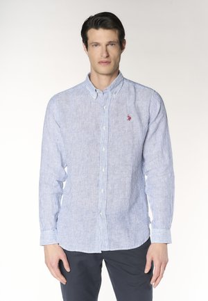 Uomo che indossa una camicia a righe azzurro chiaro e bianca con bottoni e un piccolo logo rosso sul petto, in piedi davanti a uno sfondo semplice.