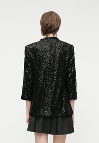 Crna jakna sa šljokicama, tričvrtin rukavima i šalom kragnom, sa teksturiranim uzorkom po cijeloj površini. Nosena preko plisirane suknje.