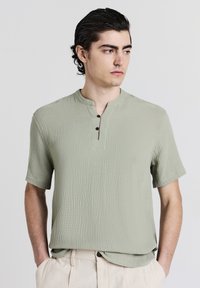 Maglietta verde a maniche corte, con texture, colletto alla cinese e tre bottoni marroni, abbinata a pantaloni beige a pieghe.