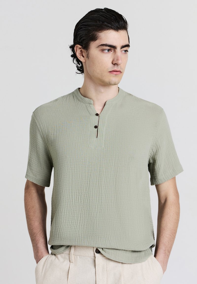 Maglietta verde a maniche corte, con texture, colletto alla cinese e tre bottoni marroni, abbinata a pantaloni beige a pieghe.