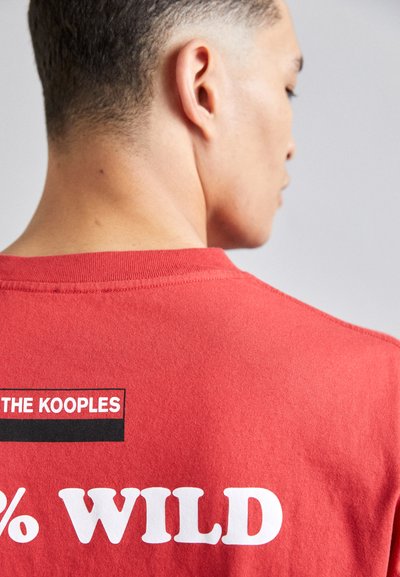 Rød bomulds T-shirt med rund hals, korte ærmer og fed hvid tekst, der siger "WILD" med "THE KOOPLES" i en sort boks.
