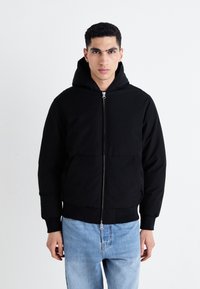 Chaqueta con capucha negra y cremallera frontal, dos bolsillos laterales, puños de canalé y una tela texturizada. Llevada con jeans de color azul claro.
