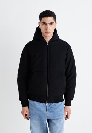 Schwarze Hoodie-Jacke mit einem Frontreißverschluss, zwei seitlichen Taschen, gerippten Bündchen und einem strukturierten Stoff. Kombiniert mit hellblauen Jeans.