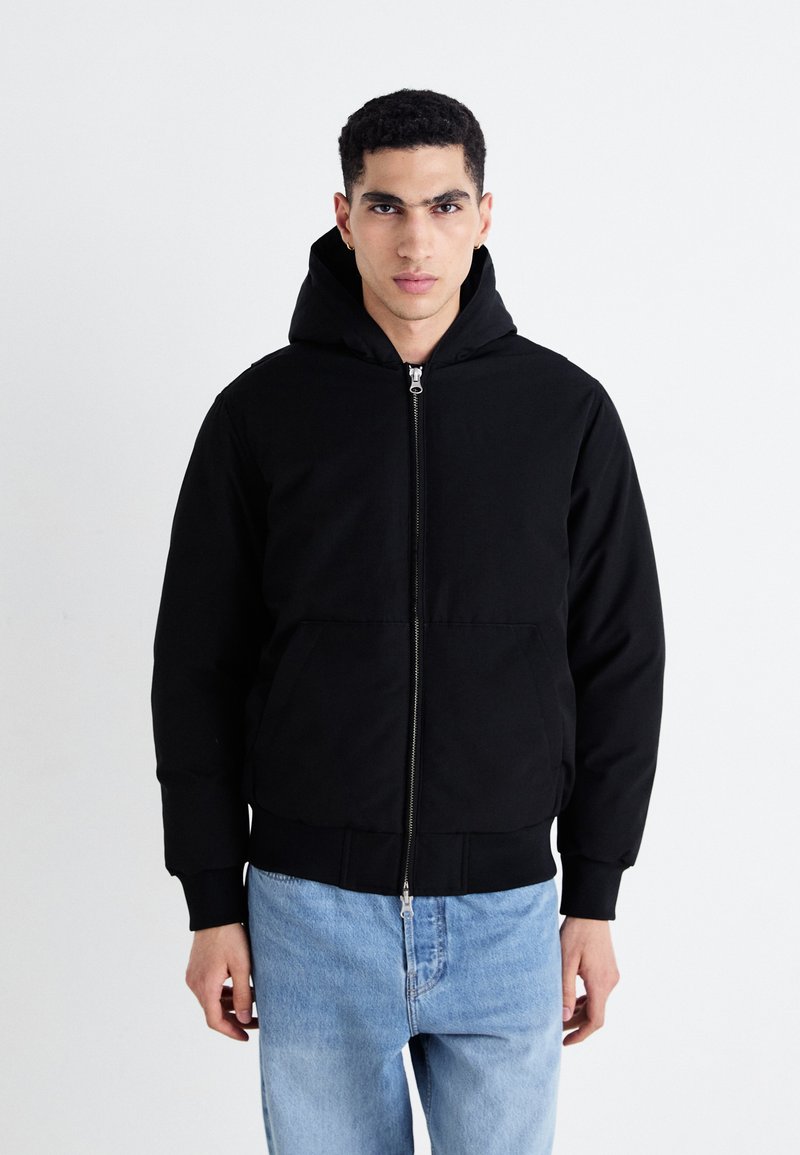 Chaqueta con capucha negra y cremallera frontal, dos bolsillos laterales, puños de canalé y una tela texturizada. Llevada con jeans de color azul claro.