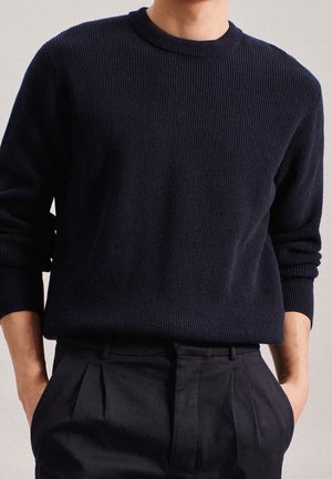 Person trägt einen dunkelblauen, gerippten Pullover mit langen Ärmeln, der in eine hoch taillierte, plissierte schwarze Hose gesteckt ist, Hände in den Taschen.