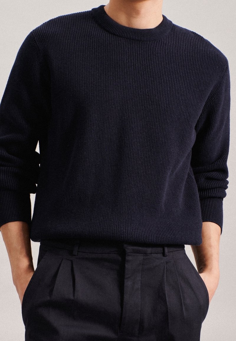 Personne portant un pull en maille côtelée bleu marine foncé à manches longues, rentré dans un pantalon noir plissé taille haute, mains dans les poches.