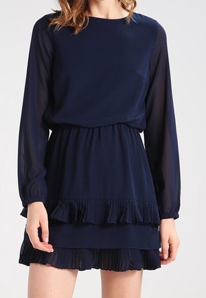 Femme portant une robe à manches longues bleu marine avec des manches transparentes et des détails à volants superposés sur la jupe.