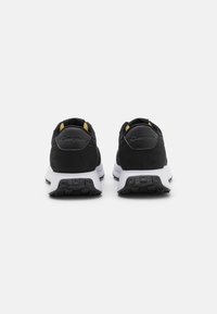 Pantofi sport negri cu un superior texturat, accente galbene și o parte intermediară albă. Vederea din spate arată un guler căptușit și un model de talpă din cauciuc.