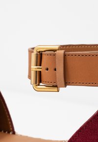 Brassard en cuir marron avec une boucle en or, attaché à une sandale en suède bordeaux. Présente des coutures le long des bords de la sangle.