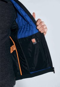 Black jacket with blue quilted lining, featuring an inner pocket, orange accents, and a zipper closure. Shell material appears smooth.