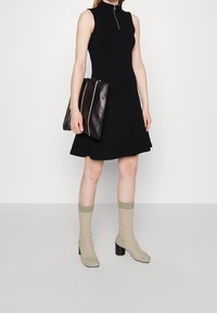 Robe noire sans manches avec un col montant et une fermeture éclair à l'avant, accompagnée d'une pochette noire avec des accents blancs et de chaussettes vertes aux chevilles avec des talons carrés.
