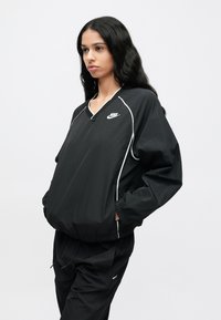 Fekete szélálló kabát V-nyakú kivágással, fehér pipinggal és Nike logóval a mellrészen. Rugalmas mandzsettákkal és bő fazonnal rendelkezik. Sima textúra.