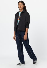 Giacca leggera nera con bottoni a pressione, indossata sopra una maglietta azzurra chiara con un logo colorato. Jeans blu scuro con cuciture a contrasto. Sneakers nere.