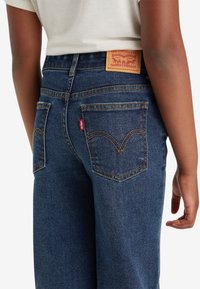 Tmavě modré džíny s vysokým pasem, s béžovou koženou nášivkou značky, žlutým prošíváním a standardními zadními kapsami s detailním logem.