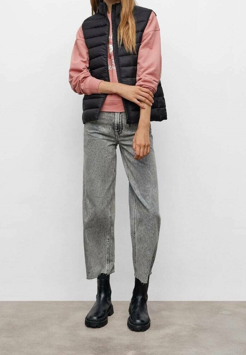 Personne portant une doudoune noire sans manches sur un sweat rose, un jean gris taille haute et des bottines noires, debout sur un sol gris.