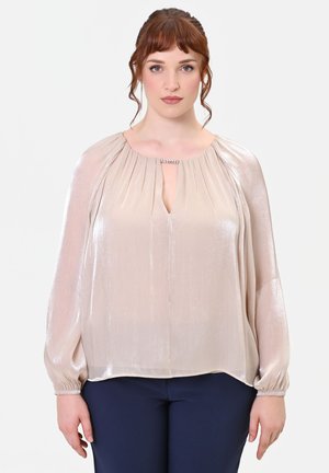 Donna con capelli rossi indossa una camicetta beige lucida a maniche lunghe con scollo decorato a forma di spacco e pantaloni blu scuro su sfondo bianco.