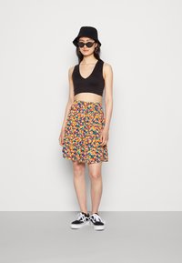 Top preto cropped, saia floral colorida com fundo azul e ténis preto e branco. Os acessórios incluem um chapéu bucket preto e óculos de sol.