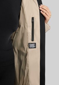 Interno beige della giacca con esterno nero, caratterizzato da una tasca laterale con zip e un'etichetta che recita "Khujo Original Changing Threads". Tessuto morbido.