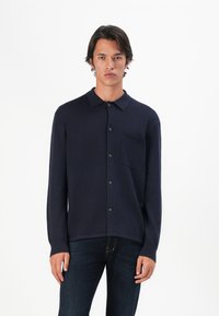 Mann in einem dunkelblauen, knöpfbaren Langarmhemd mit Brusttasche und dunklen Jeans, der vor einem schlichten weißen Hintergrund posiert.