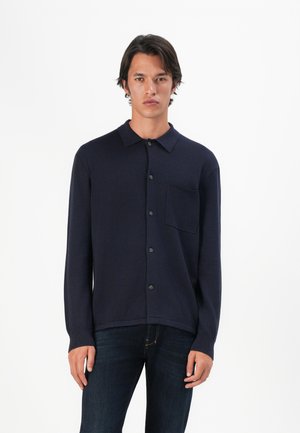 Mann in einem dunkelblauen, knöpfbaren Langarmhemd mit Brusttasche und dunklen Jeans, der vor einem schlichten weißen Hintergrund posiert.