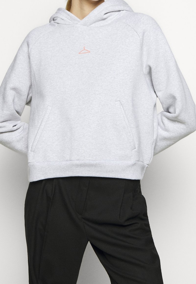 Personne portant un sweat à capuche gris clair avec une petite icône de cintre orange sur la poitrine et un pantalon noir, mains dans le dos.