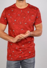 Man i röd kortärmad t-shirt med små racingsymboler och blå jeans, händerna hopknäppta framme, mot en enkel ljus bakgrund.