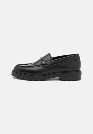 Mocassin en cuir noir avec un bout arrondi, un dessus élastique et une semelle en caoutchouc texturée. Présente un détail décoratif en métal argenté.