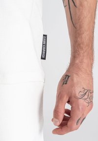 Material blanco texturizado con un diseño ajustado, que presenta una etiqueta negra con "Gianni Kavanagh". Primer plano de una mano mostrando tatuajes.