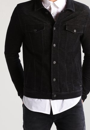 Mann trägt eine eng anliegende schwarze Jeansjacke über einem weißen Hemd mit Knöpfen, die Hände in den Taschen, kein sichtbarer Kopf oder Füße.