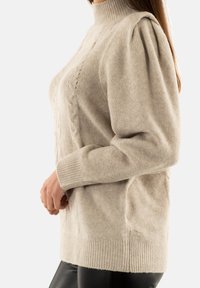 Maglione beige lavorato a maglia con collo alto, maniche a palloncino, polsini a coste e un motivo testurizzato sul davanti. Realizzato in materiale morbido e caldo.