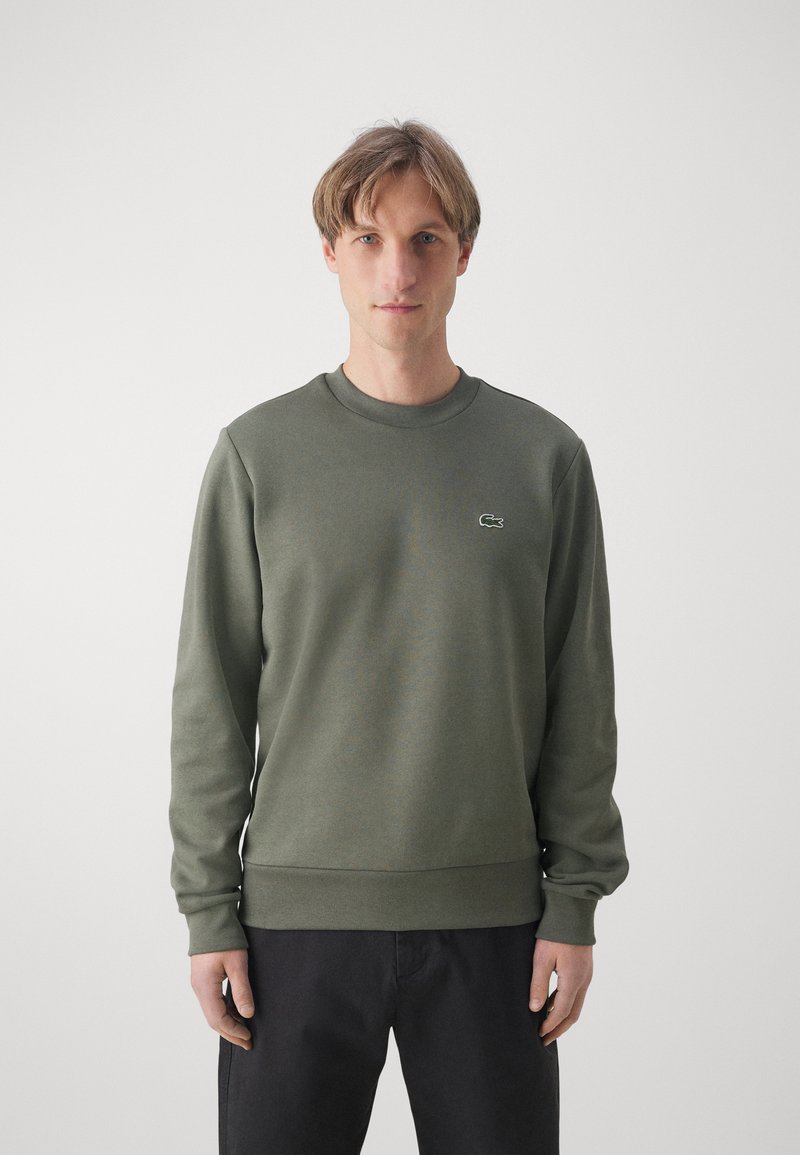 Giovane uomo che indossa un maglione a girocollo verde oliva con un piccolo logo di coccodrillo sul petto, in piedi contro uno sfondo chiaro e semplice.