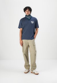 Mörkblå bomull T-shirt med grafiskt tryck, beige cargobyxor med flera fickor, och vita sneakers med detaljer. Scarf i blått mönster.