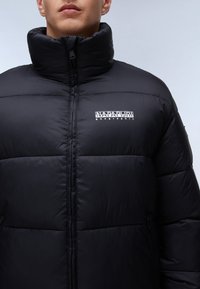 Svart puffjacka med hög krage, dragkedjestängning och framsidan med logotyp. Slät, vadderad textur visar segmenterade paneler och sömmar.