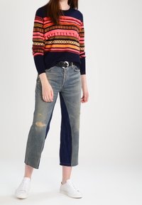 Femme portant un pull rayé multicolore, un jean court bicolore avec des détails usés, une ceinture noire et des baskets blanches, debout sur un fond blanc.