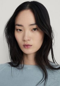 Primo piano di una persona con lunghi capelli neri, che indossa una maglietta di tessuto azzurro chiaro, con un tono di pelle naturale e un'espressione facciale neutra.
