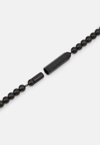Svart perleaccessoire laget av glatt materiale, med en rektangulær kobling med preget detaljer. Generelt slank og matt finish.