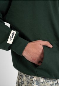 Personne portant un sweat-shirt vert foncé avec un patch brodé sur la manche, mettant la main dans la poche, montrant un petit tatouage rouge sur la main au-dessus du poignet.