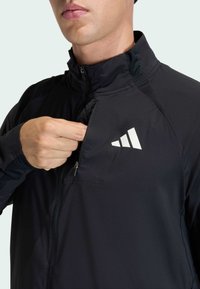 Black zip-up jacket featuring a high collar, lightweight material, and a small zippered pocket on the chest with a white logo accent.