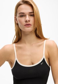 Crni top na bretele s kremasto obojenim trakama i malim izvezenim logotipom. Glatka tekstura, pripijeni kroj.