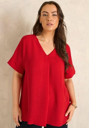 Kvinde med langt brunt hår iført en løs rød kortærmet V-udskæringsbluse, stående foran en ensfarvet lys beige baggrund.