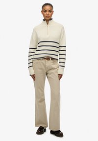 Suéter a rayas crema y marino con cuello de cremallera, combinado con pantalones de pierna ancha beige y zapatos marrones. Textura suave y ajuste relajado.