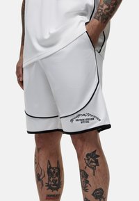 Pantaloni corti sportivi bianchi in rete, con dettagli neri, piping e logo ricamato. Tatuaggi visibili sulle gambe.